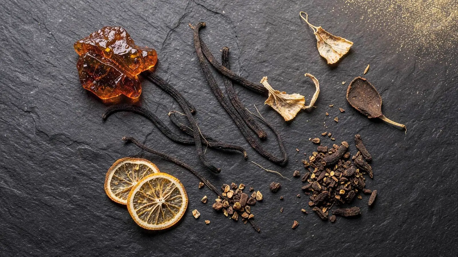 Flat lay of amber resin, vanilla pods, citrus peel and spices on dark slate