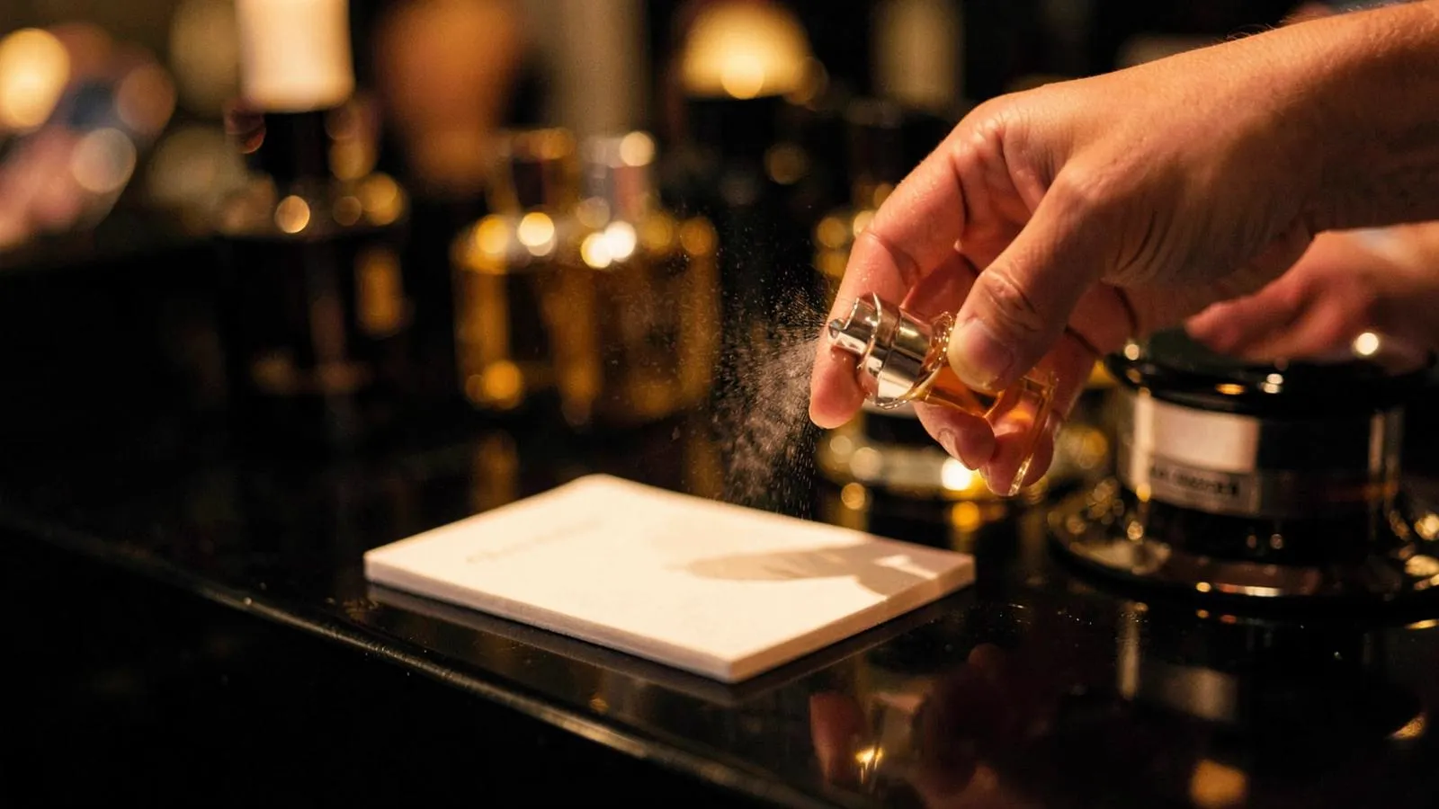 A hand spraying perfume onto a blotter strip beside testers on a dark counter